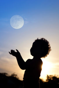 A young boy looks up at a full moon as the sun sets.