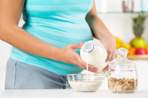 A pregnant woman pours milk into a bowl of cereal in a kitchen.