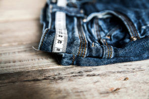 A pair of jeans is in focus. Measuring tape is looped through the jean's belt loops.