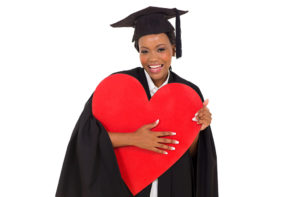 A woman wears a graduation cap and gown while holding a heart-shaped object.