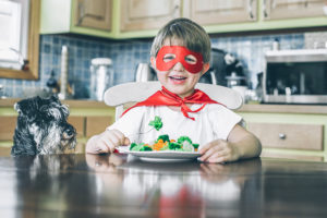 A young boy wears a red superhero mask and cape as he eats broccoli at a kitchen table.