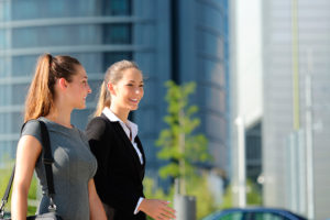 Two women walk outside together. They wear business casual clothing.