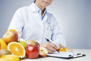 A medical professional sits at a table and writes on a clipboard. The table holds measuring tape and fruit.