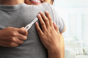 A woman hugs a man. The woman holds a pregnancy test in her hand.