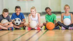 A group of kids sit on the floor and hold different kinds of balls. Basketballs, socerballs and volleyballs are shown in the kids' hands.