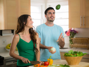 A woman cuts up bell peppers as a man juggles an avocado and onion.