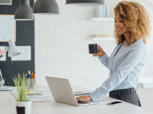 A woman works on her laptop as she stands and drinks coffee.