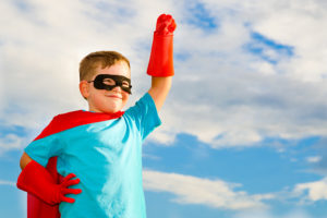 A young boy wears a superhero cape, gloves and a mask outside. He raises his hand in the air like he is flying.