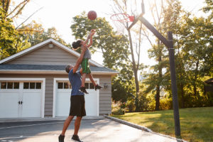 A father lifts his child up to shoot a basketball into a hoop outside.
