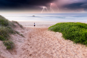 A person stands on the beach and looks into the distance. A storm cloud is coming toward them with lighting shown in the background.