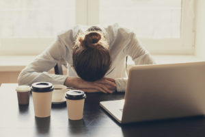 A woman lies her head on a table to take a nap. The table contains multiple cups of coffee and a laptop.