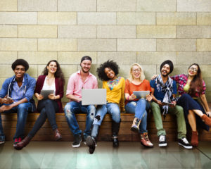 A group of college students pose for a photo together. They sit on a long bench in front of a brick wall.