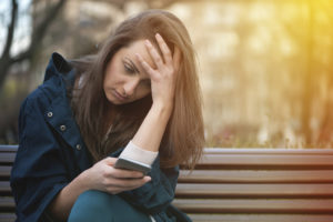 A woman sits on a bench and stares at her phone. She rests her head against her hand and appears upset.