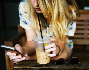 A young woman holds an iced coffee in one hand and her cellphone in the other hand.