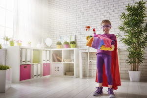 A young girl holds cleaning products as she wears a superhero cape and mask.