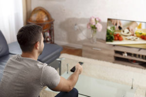 A man points his TV remote toward a TV. The man watches a cooking show.