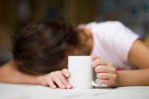 A person rests their head on their desk. Their hands are holding a cup of coffee.