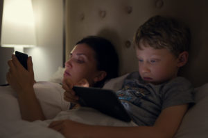 A mother and her son lie in bed and watch something on their portable, electronic devices.