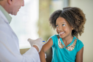 A brave little girl is getting a flu shot from a doctor. He is holding the needle and is about to give her the shot.