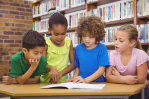 A group of kids stand over a table and read a book. 