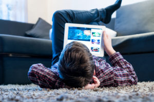 A young boy lies on the floor and stares at a video on his electronic tablet.