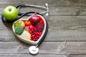 A heart-shaped bowl holds fruits and vegetables. A green apple and stethoscope is plaved next to the bowl.
