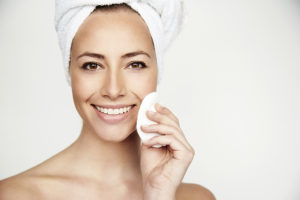 A woman wears a towel around her hair and washes her face. She smiles while holding a cotton pad.