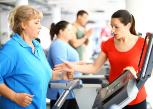 A woman wearing a blue shirt walks on a treadmill at a gym. A woman wearing a red shirt appears to be talking to her.