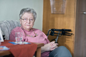 An elderly adult sits in a chair. Her side table contains a glass of water.
