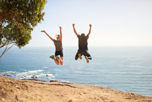 Two people jump off a cliff with their hands in the air into a body of water.