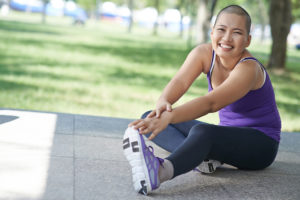 A bald woman stretches out her legs and smiles. She is wearing a purple tank top and purple tennis shoes.
