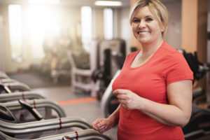 A woman walks on a treadmill and smiles. She wears a red T-shirt and is at the gym.