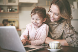 A woman sits with a young child and watches something on a laptop. The child is laughing.