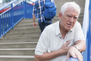 An elderly man leans against a stair railing. He places his hand over his heart and appears in pain.