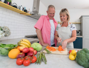 A woman wears an apron and cuts up an orange bell pepper in a kitchen. A man looks on. The kitchen counter is filled with fruits and vegetables.