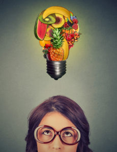 A woman looks up at a lightbulb. The lightbulb is made out of fruit.