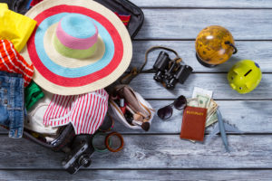 A suitcase is shown with a sun hat, jeans, swim suit, sunglasses, binoculars, makeup, a camera, pony tails, a passport and much more.