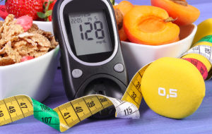 A black blood sugar monitor reads, 128. The black monitor is placed next to fruit, cereal, measuring tape and a yellow 0.5 dumbbell.