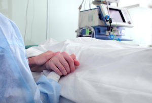A person lies in a patient bed while a loved one holds their hand.