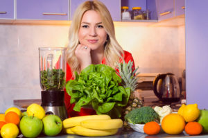 A woman poses for a photo next to her juicer, fruits and vegetables.