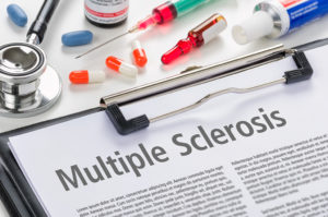A clipboard with a piece of paper reading, "Multiple Sclerosis" is in focus. Next to the clipboard there are pills, medications and a stethoscope.