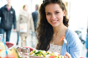 A teenage girl sits a table and eats a salad. She smiles.