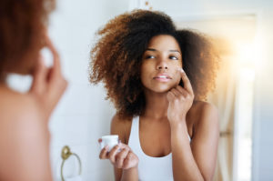 A woman stands in front of a mirror in her bathroom and applies a skin-care product on her face with her finger.