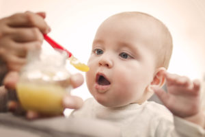 A baby opens its' mouth as a person feeds them yellow-colored baby food.