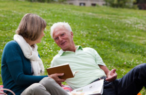 Two elderly adults lie in the grass and read a book. They appear to be talking about the book they are reading.