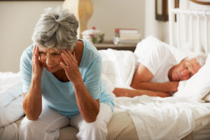 A elderly woman sits on her bed placing her hands on her forehead. Her partner appears asleep in bed. She is wide awake and upset. 