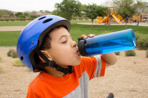 A young boy stops riding his bike to take a sip of water from his blue water bottle. The boy wears a blue helmet.