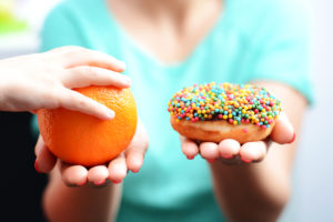 A woman holds a donut in one hand and an orange in another. A person places their hand on top of the orange.