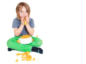A young kid stuffs his mouth with orange-colored cheese puffs.