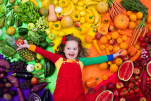 A young girl lies on a floor covered with fruits and vegetables. The fruits and vegetables are separated by color.